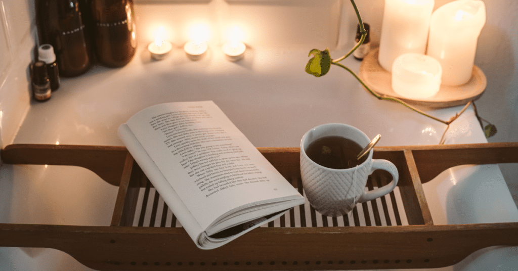 A book and hot tea are set on a bath tray, prepared to be enjoyed during a hot bath surrounded by candles and plants _ Self care activities for adults