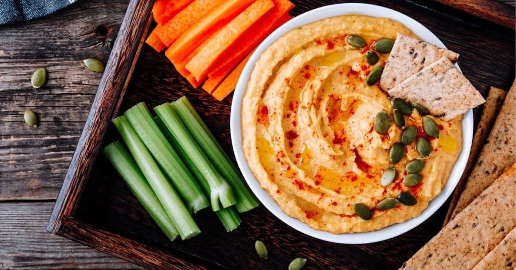 Hummus sits in a white bowl on a wooden tray with celery sticks, carrot sticks, and seeded crackers | Hummus and Misleading Food Labels