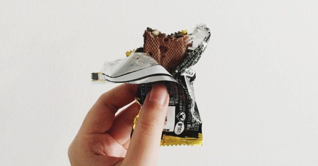 A woman's hand holds up a protein bar with the wrapper ripped partially off showing a chocolate covered protein bar with a bite taken out of it