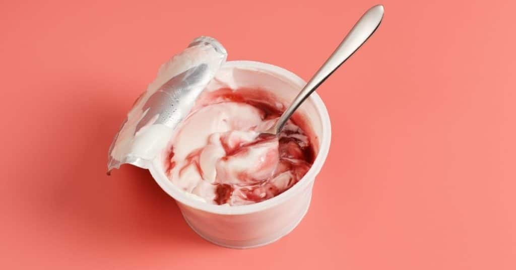 An unlabeled yogurt container has the foil lid partially peeled off with a spoon inside showing premixed fruit flavoring added to the yogurt | Misleading Food Labels