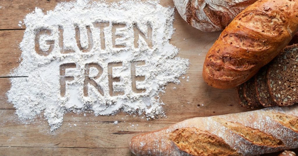 A pile of flour is on a wooden countertop surrounded by loaves of fresh baked bread. In the flour is written the words "Gluten Free"