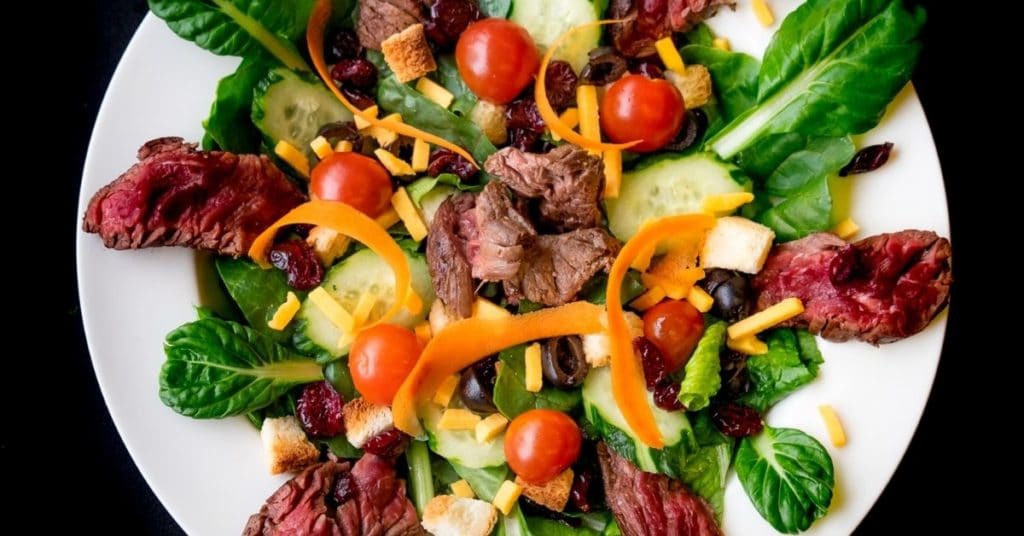 A birds-eye-view of a dark leafy green salad, topped with tomatoes, cheddar cheese, carrot strips, cucumber, and slices of cooked steak, showcasing a great gluten free choice for those with iron deficiency and gluten intolerance