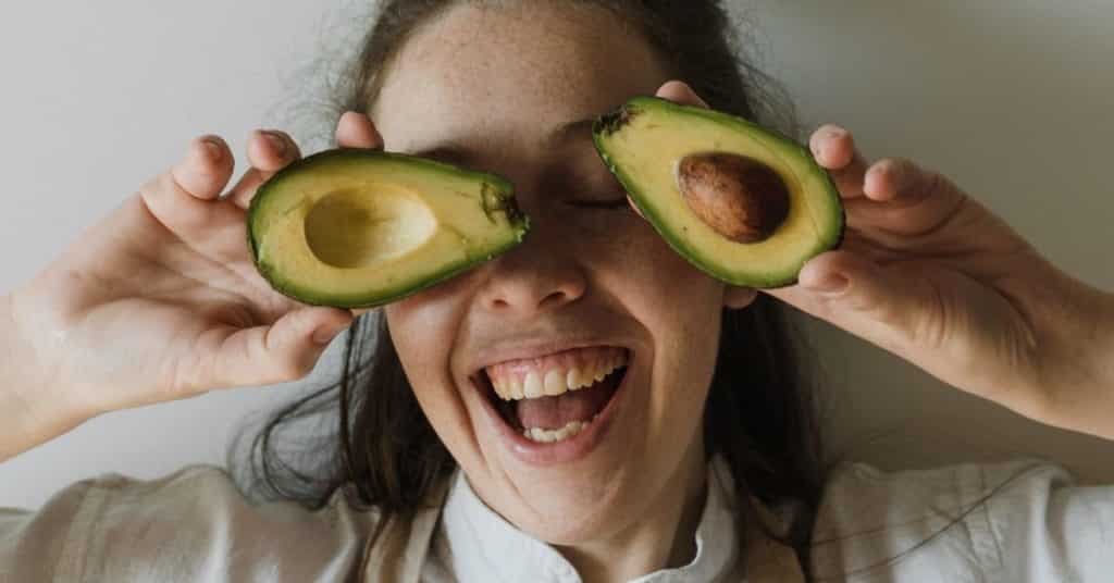Avocado Benefits for Health, A woman smiles with an open mouth as she holds up a halved avocado, with each half covering one of her eyes