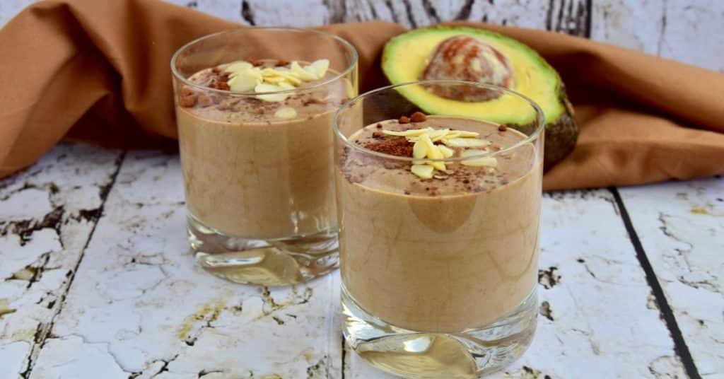 Two chocolate smoothies in glasses, topped with cocoa powder and almond slivers are placed in front of a half of an avocado and a decorative cloth