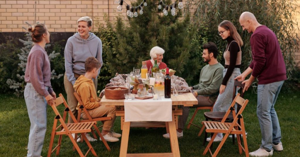 Outdoor Easy Summer Dinner Party Menu, A family stands around an outdoor table and seating chatting and smiling with a bunch of food and drinks spread out on the table top