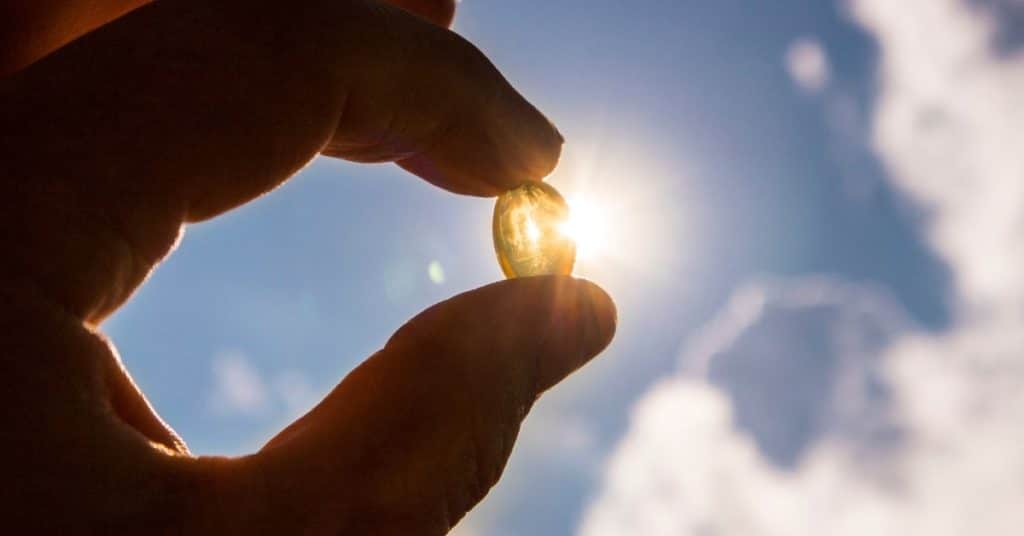 A hand holds up a semi-transparent gel Vitamin-D supplement pill to the sun, the light from the sun bursts through the pill like a glowing gem