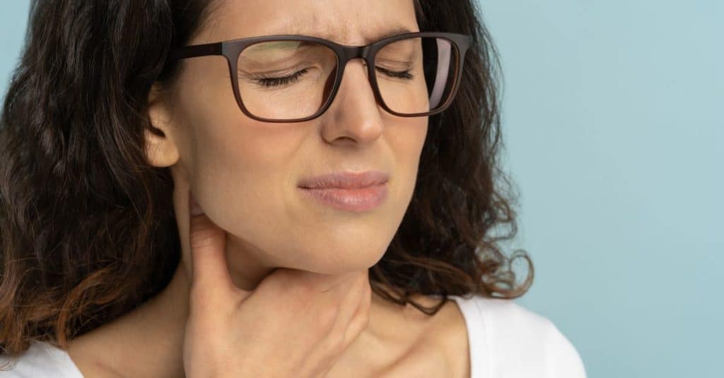 A dark-haired woman in glasses looks in pain and holds her throat from acid reflux, How to Increase Your Stomach Acid Naturally