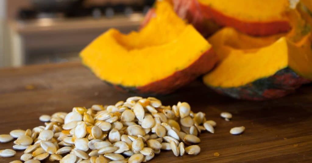 Pumpkin, a seasonal fall food, diced into large chunks on a wooden cutting board with all of the seeds scooped out and into a pile in front of the fruit