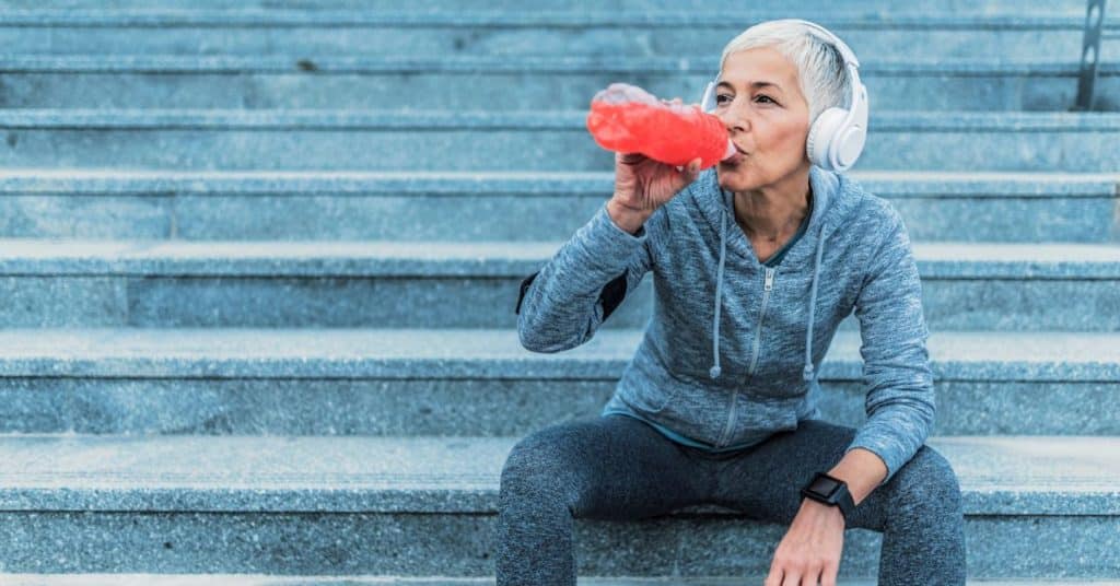 A gray-haired woman in headphones, smartwatch, and athletic sweatshirt and leggings sits on bleachers and sips a water bottle with electrolytes in it