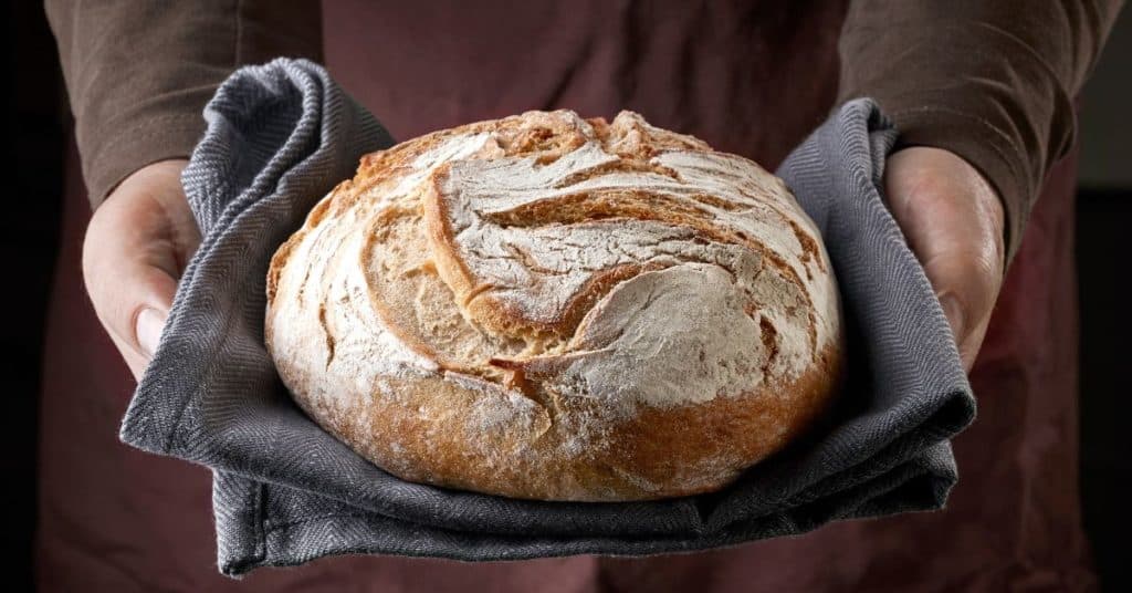 A person's hands hold out a fresh loaf of sourdough bread nestled in a kitchen towel
