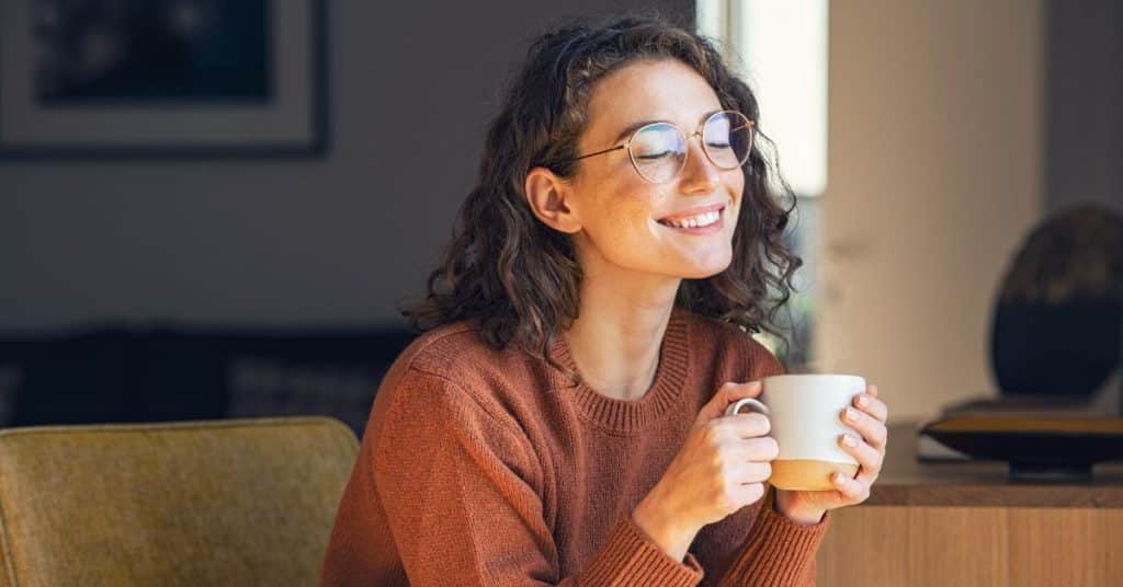 A young woman in glasses sits on a couch and happily sips a mug of hot tea, hydrate with hot beverages in the winter