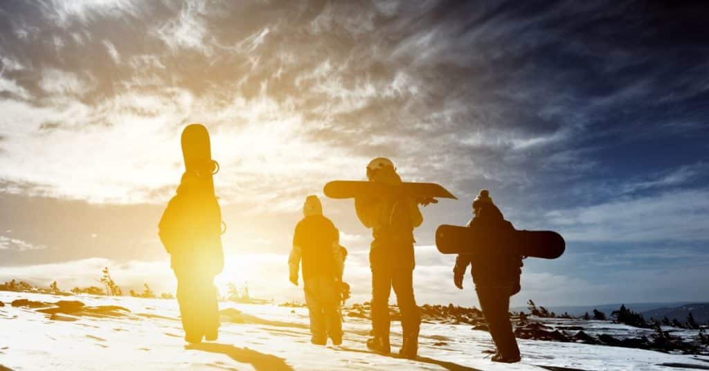 Four people's silhouettes, each carrying snowboards, hike up a snowy hill, hydrate after winter exercise