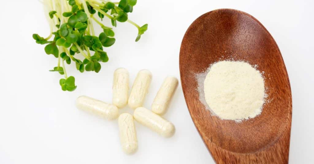 A wooden spoon with a fine powder is displayed next to multiple pill capsules and fresh broccoli sprouts, What foods remove toxins from your body