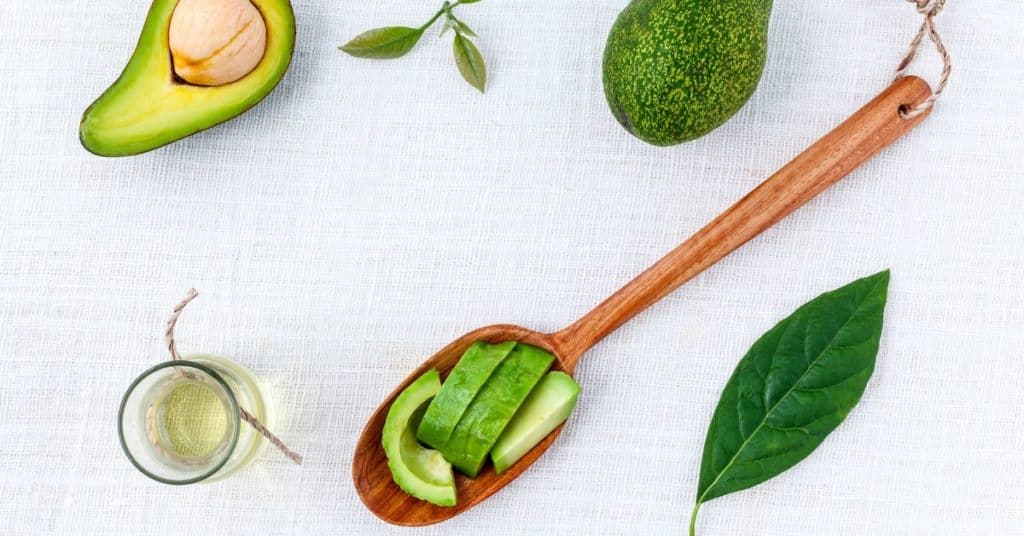 Halved avocados and diced avocado are displayed with a wooden spoon and carafe of avocado oil