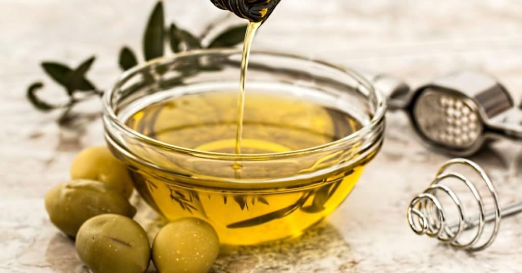 Olive oil is poured from an opaque bottle into a small glass bowl surrounded with olives and olive leaves