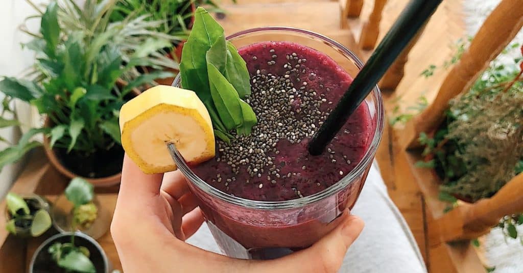 A woman's hand holds out a purple smoothie garnished with green herbs, chia seeds and a banana slice