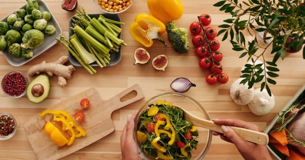 A woman's hands mix up a salad, her chopping block is scattered with colorful fruits and vegetables like peppers, garlic, figs, avocado, tomatoes, asparagus, Brussels sprouts and more