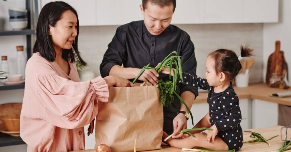 A mom and dad unpack a grocery bag while their little one plays with the produce on the kitchen counter