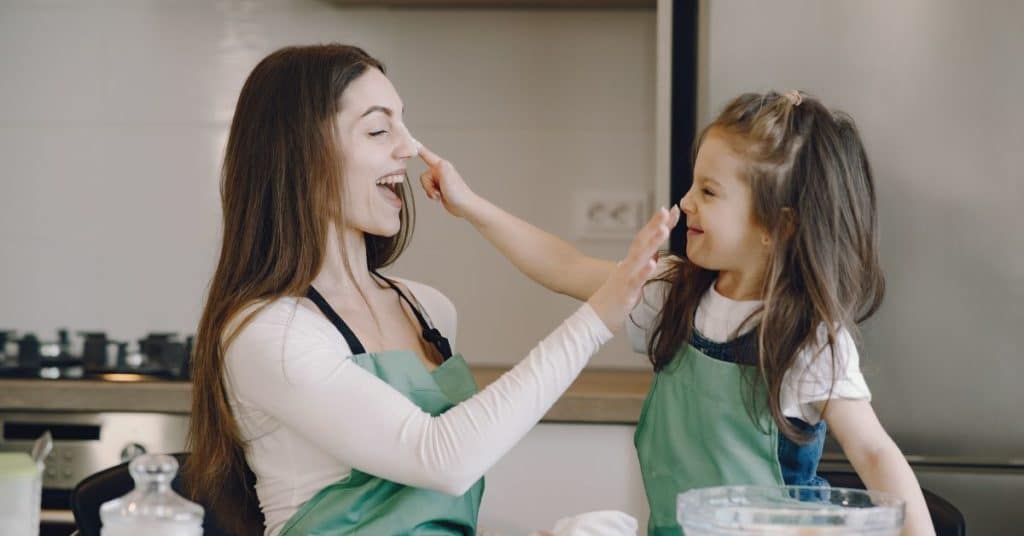 A mom and daughter playfully rub flour on each other's noses while they bake together in the kitchen
