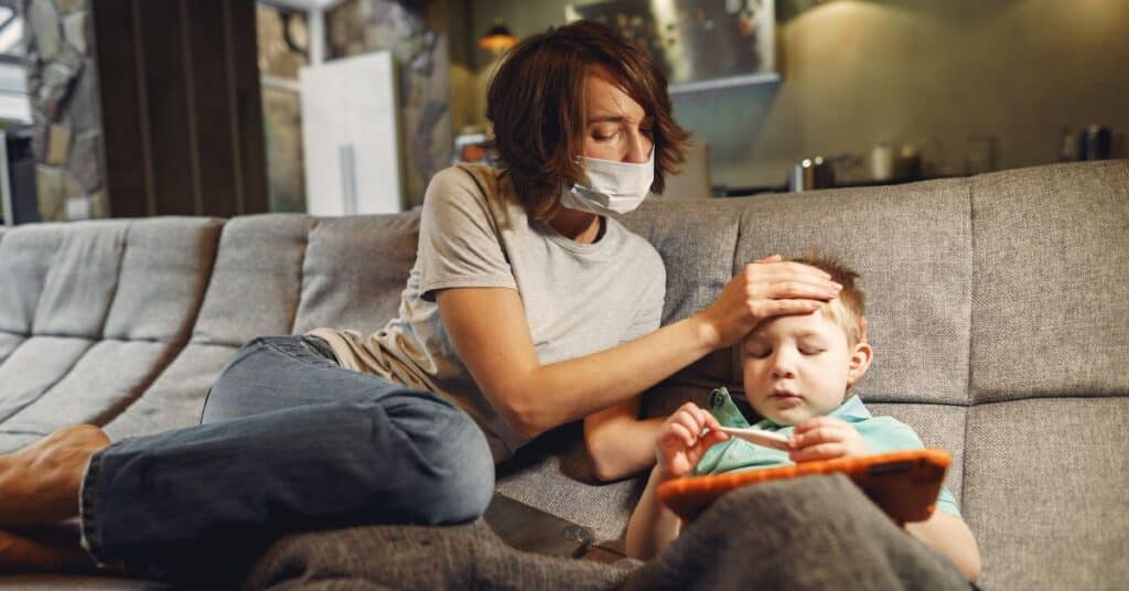 Mom in face mask sits on couch with her son and checks for a fever while he plays on a tablet, Child complains of stomach pain every day