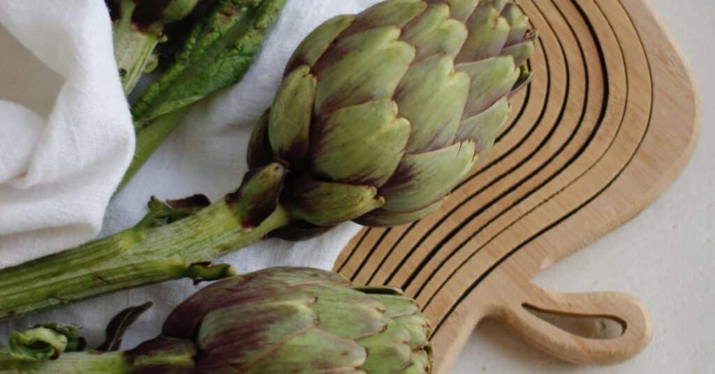 A full ripe artichoke bud rests on a clean tea towel and wooden cutting board, recipes to help detox
