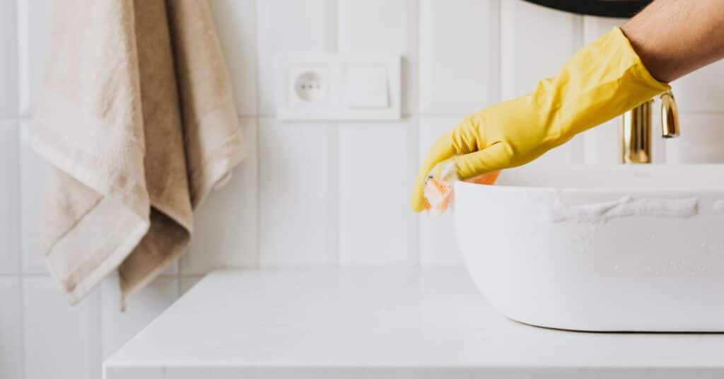 A hand covered in a rubber glove wipes a sink with a soapy sponge