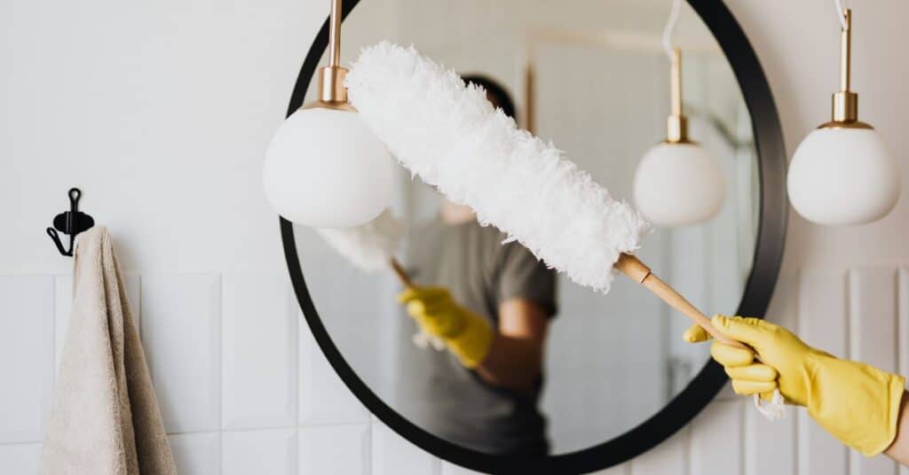 A hand in rubber glove holds up a microfiber dusting wand in front of a bathroom mirror as it reaches for the lights to dust them