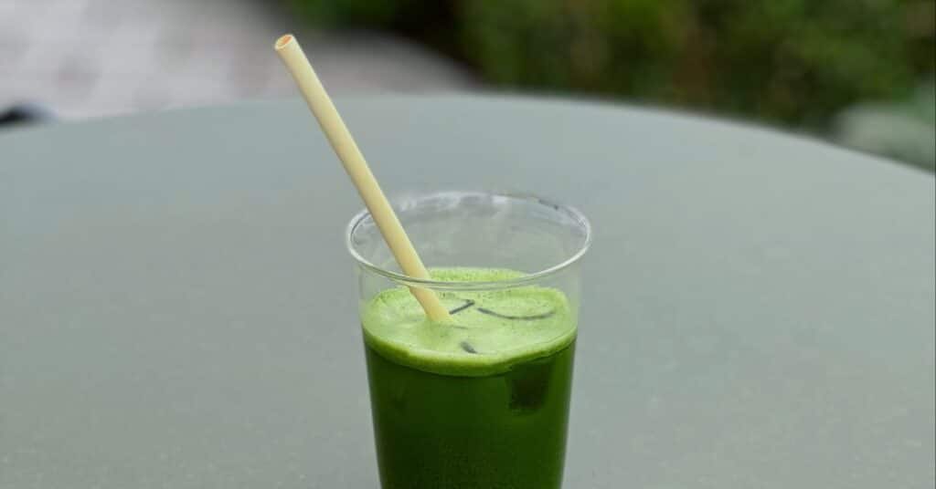 A healthy looking green chlorella drink in an opaque glass with a drinking straw, ice cubes and slightly frothed top sits on an outdoor dining table, foods to help detox