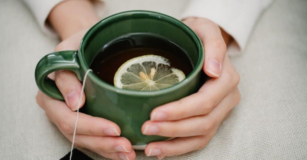 A woman's hands hug a large ceramic mug with hot liquid, a tea bag and slice of lemon in it, foods to help detox