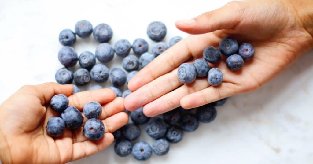 Two hands hold large blueberries above a countertop scattered with more berries, foods to help detox