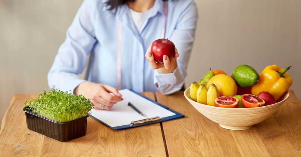 A registered dietitian holds out an apple with a clipboard and bowl of fruit in front of her, Registered Dietitian vs Nutritionist