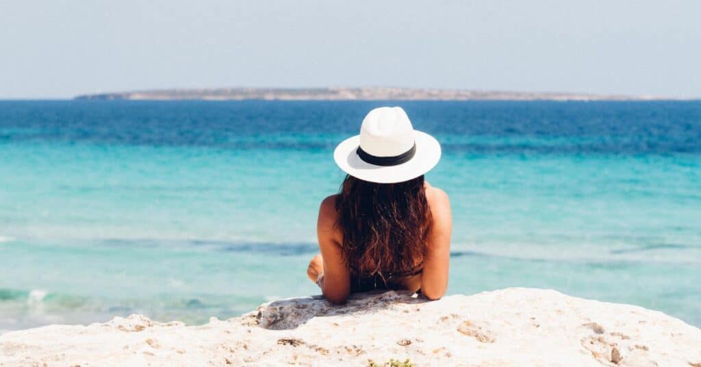 A woman in a white fedora leans on a rock and looks out at the ocean in the summer sun