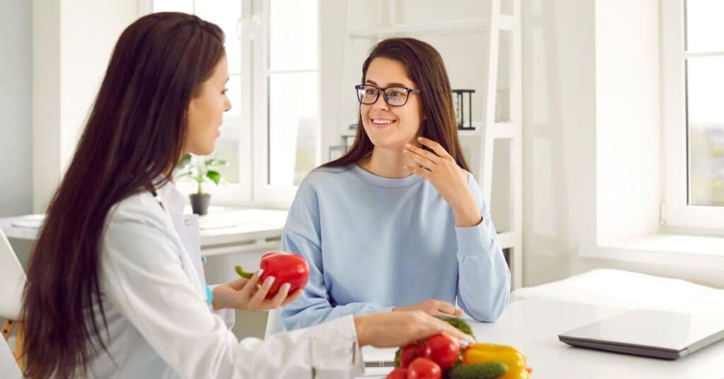 A woman talks to a registered dietitian who is holding up various fruits and vegetables, Registered Dietitian vs Nutritionist