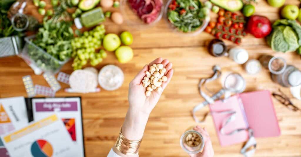 A woman's hand holds out a handful of cashews above a table full of nutrition charts and various foods, Registered Dietitian vs Nutritionist