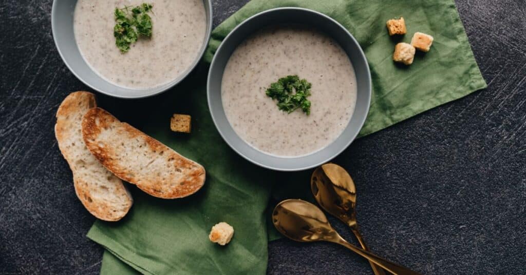 A spiced creamy soup garnished with parsley sits in two bowls, the scene is finished off with bits of toasted bread, spoons and cloth napkins