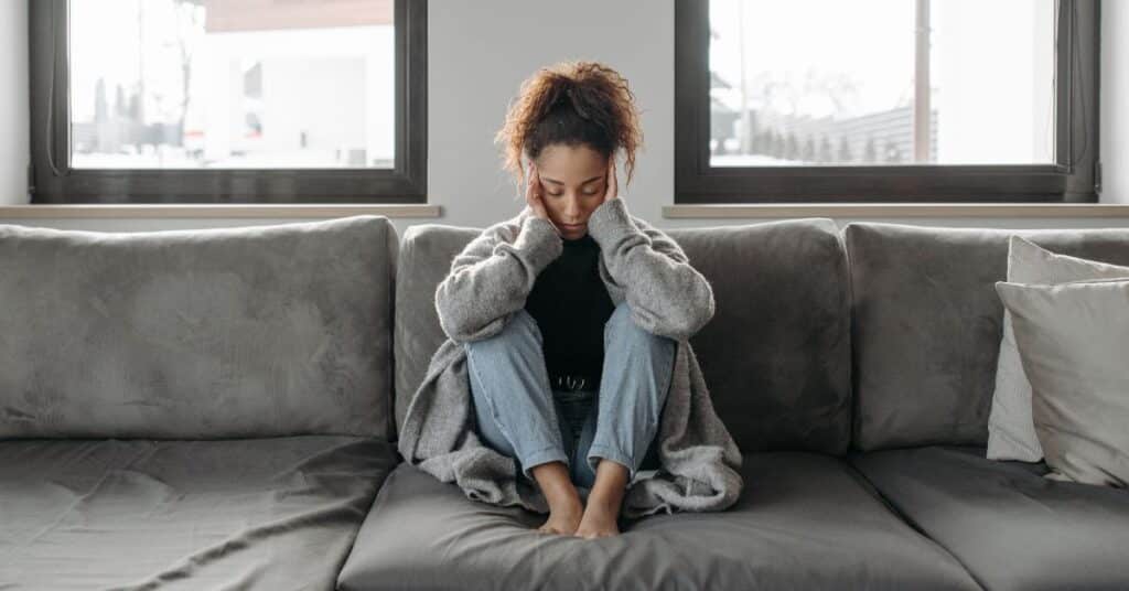 Woman sitting on a gray couch holding her head between her hands with closed eyes, food sensitivity headache