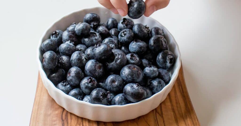 A hand grabbing a blueberry out of a bowl of antioxidant-rich blueberries sitting on a wooden cutting board, How to Lower Insulin Quickly