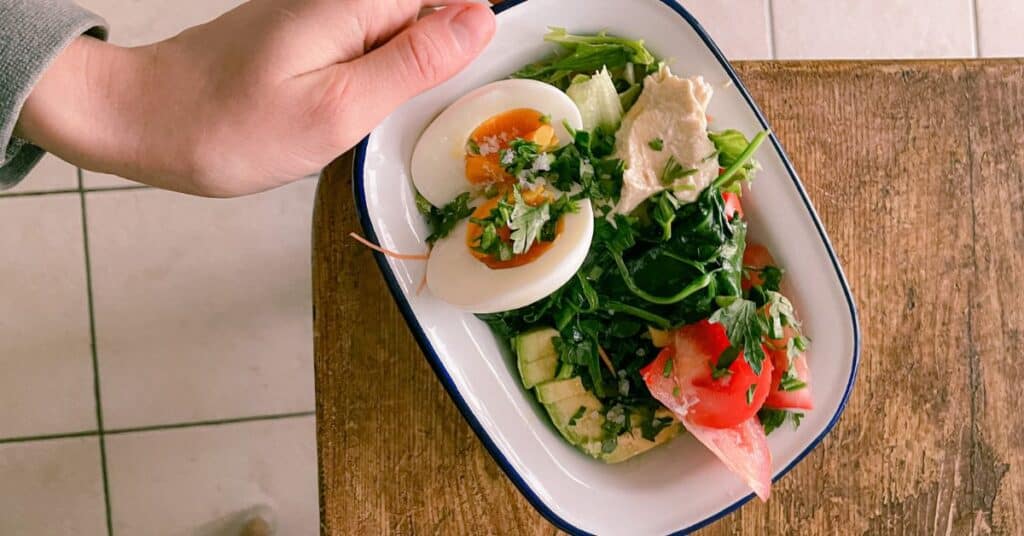 Hand holding a bowl with eggs, avocado, lunch meat and greens