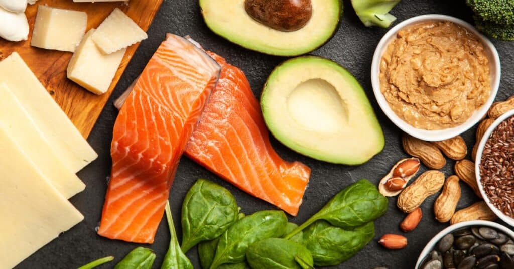 Keto foods on a black table includeing cheese, salmon, avocado, spinach and peanut butter, keto diet advantages and disadvantages