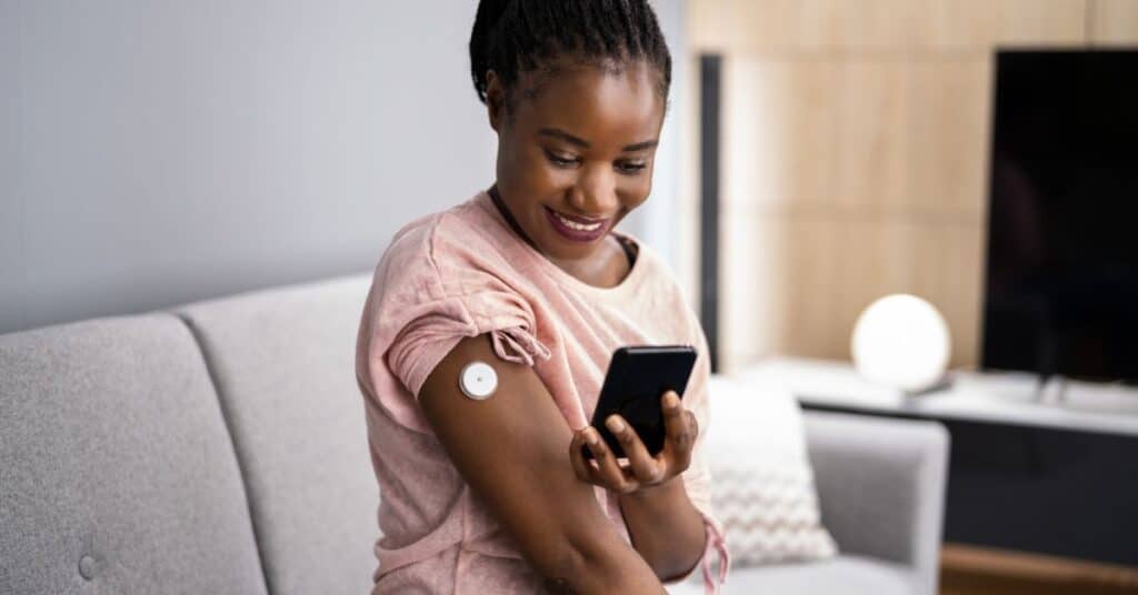 Smiling woman holding her phone in hand with a continuous blood glucose monitor in her upper arm, benefits of continuous glucose monitoring