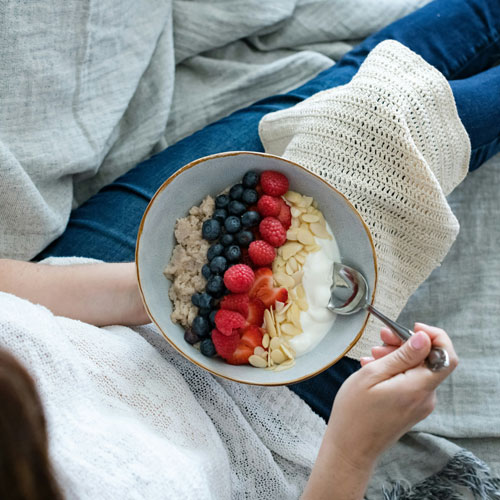 Woman sits on her couch and eats a berry, yogurt and granola bowl, anti-inflammatory foods