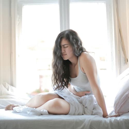 Woman sits up in her bed and clutches her stomach in pain